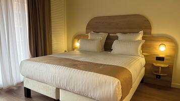 A neatly made bed with a wooden headboard, two bedside lamps, and a large window with curtains.