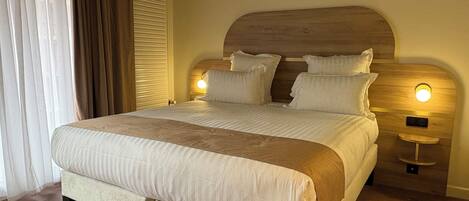 A neatly made bed with a wooden headboard, two bedside lamps, and a large window with curtains.