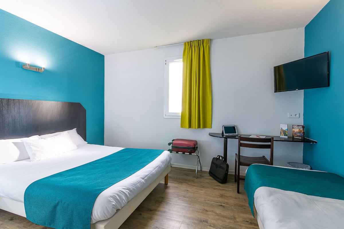 Une chambre d’hôtel avec un grand lit, un lit plus petit, un bureau avec une chaise, un téléviseur fixé au mur et une fenêtre avec des rideaux.
