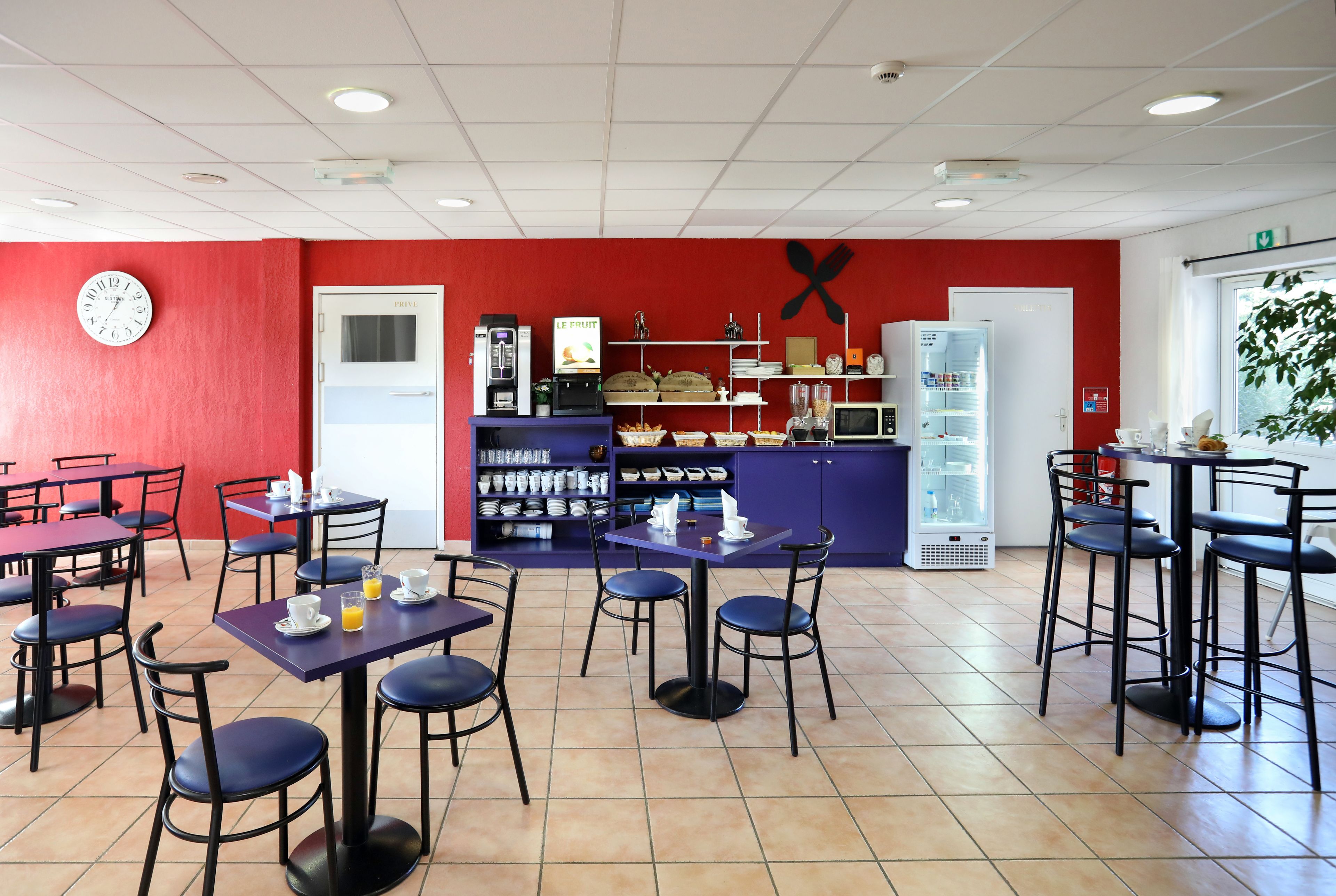 Un café con paredes rojas, varias mesas y sillas, un reloj y diferentes mostradores con comida y bebidas.