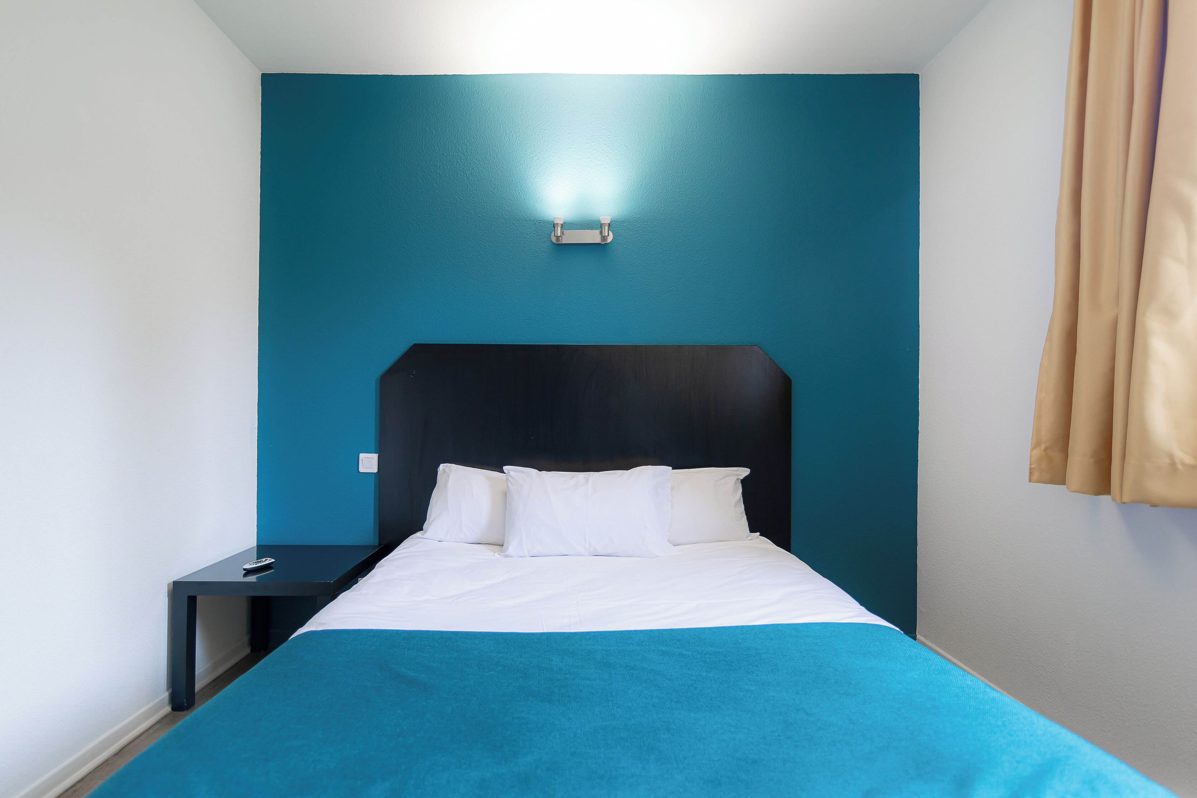 Una cama bien hecha con ropa blanca y un cabecero azul verdoso en una habitación con paredes azul verdosas y una mesita de noche pequeña.