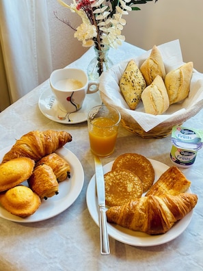 Mesa de café da manhã com croissants, panquecas, um copo de suco de laranja, uma xícara de café e uma cesta de pão.