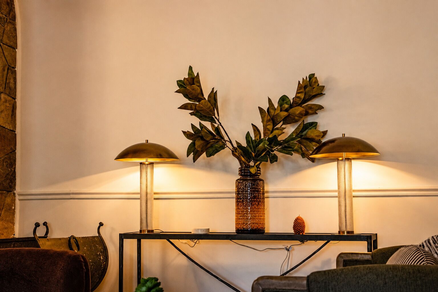 A room with a console table, two lamps, a decorative vase with foliage, and a stone wall.