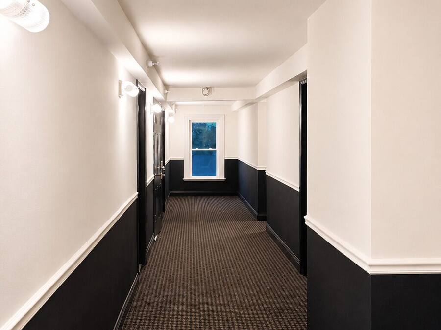 Um corredor de hotel com piso coberto por carpete, paredes brancas e detalhes em preto.