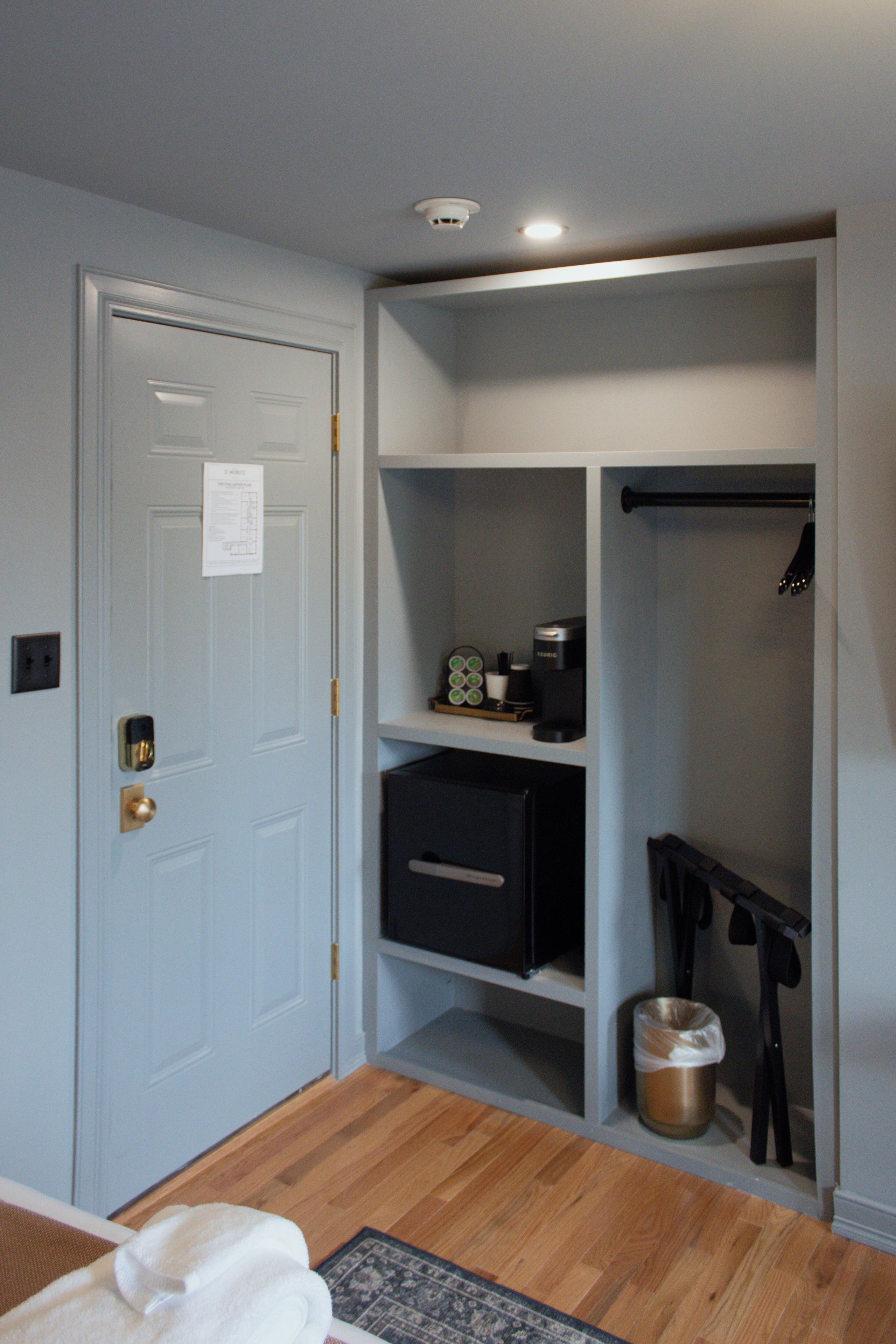 A room with a closed door, a coffee maker on a shelf, a trash can, and a coat rack.