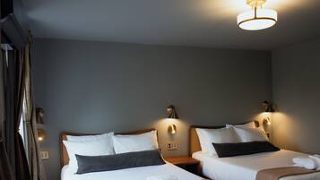 Une chambre d’hôtel avec deux lits, chacun doté de draps blancs et d’oreillers foncés, une table de chevet, une lampe fixée au mur et un rideau.