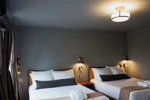 A hotel room with two beds, each with white linens and dark pillows, a bedside table, a wall-mounted lamp, and a curtain. - Hotel St. Moritz (Lake Placid)