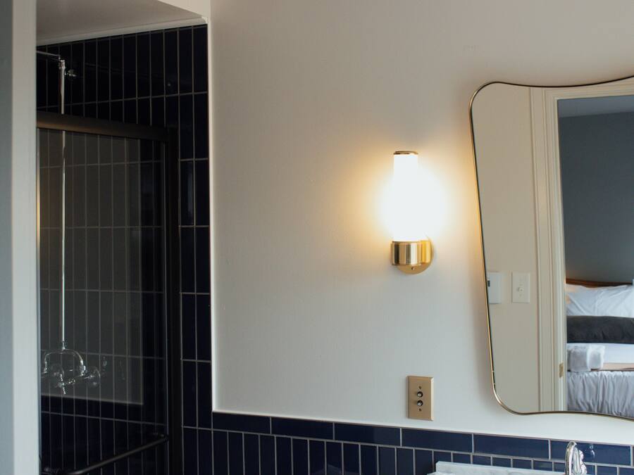 Ett hotellbadrum med dusch, en väggmonterad lampa och en spegel som reflekterar en säng.