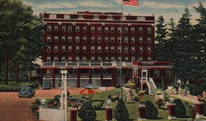 A multi-story hotel with a red facade, a flag, and a front porch with outdoor seating. - Hotel St. Moritz (Lake Placid)