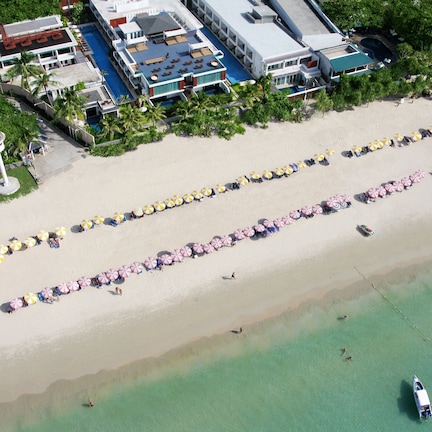 An aerial view of a resort with multiple buildings, a beach with rows of umbrellas, and a clear blue ocean.
