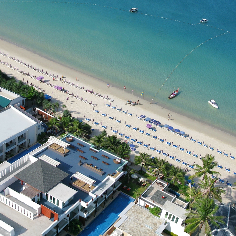 An aerial view of a beach resort with a long stretch of beach, multiple boats, and a pool area with sun loungers.
