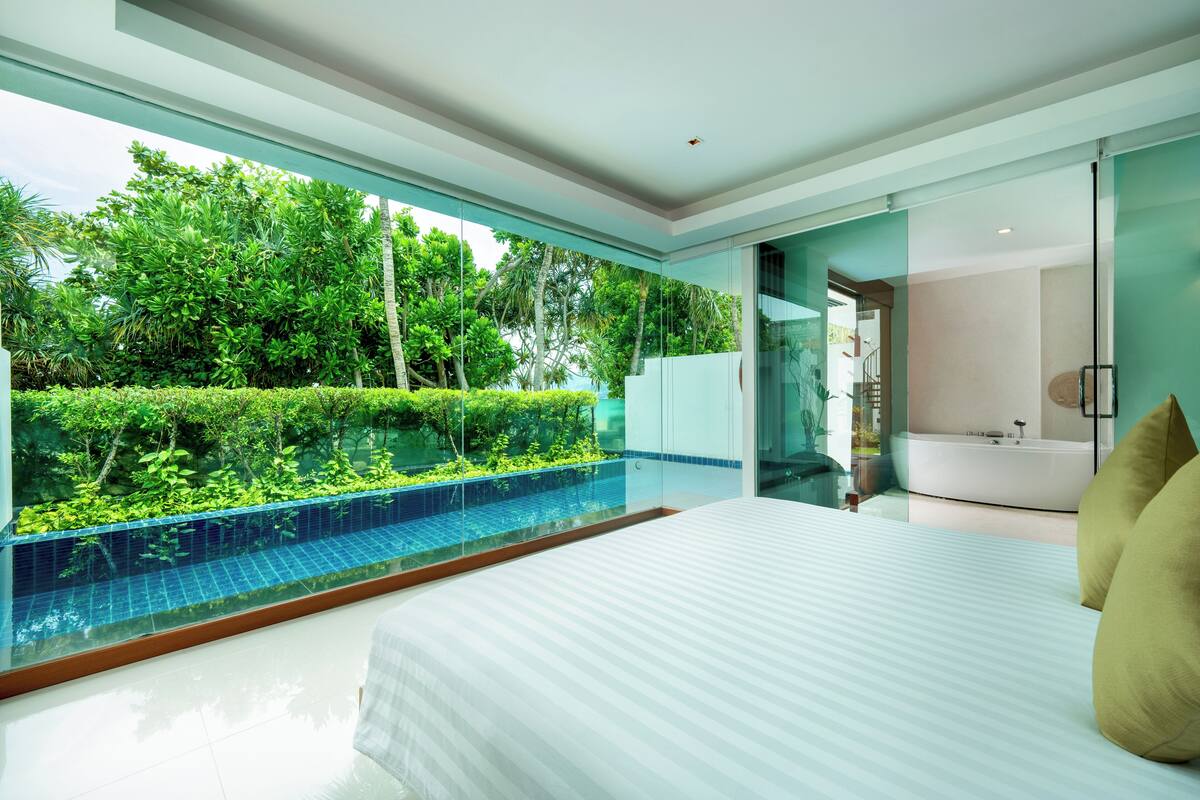 A modern bedroom with a large bed, a glass wall overlooking a pool and lush greenery, and a separate bathroom with a bathtub.