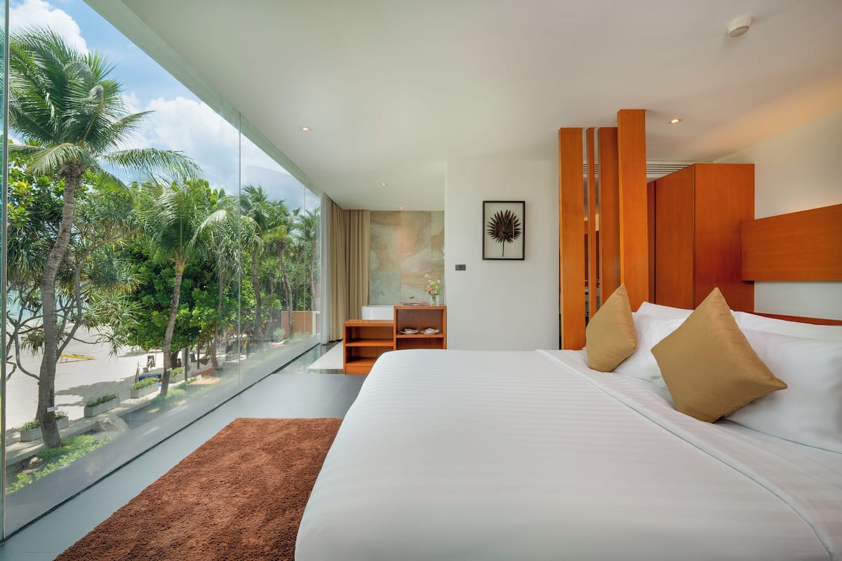 A modern hotel room with a large bed, a wooden wardrobe, a nightstand, and a view of palm trees through large windows.