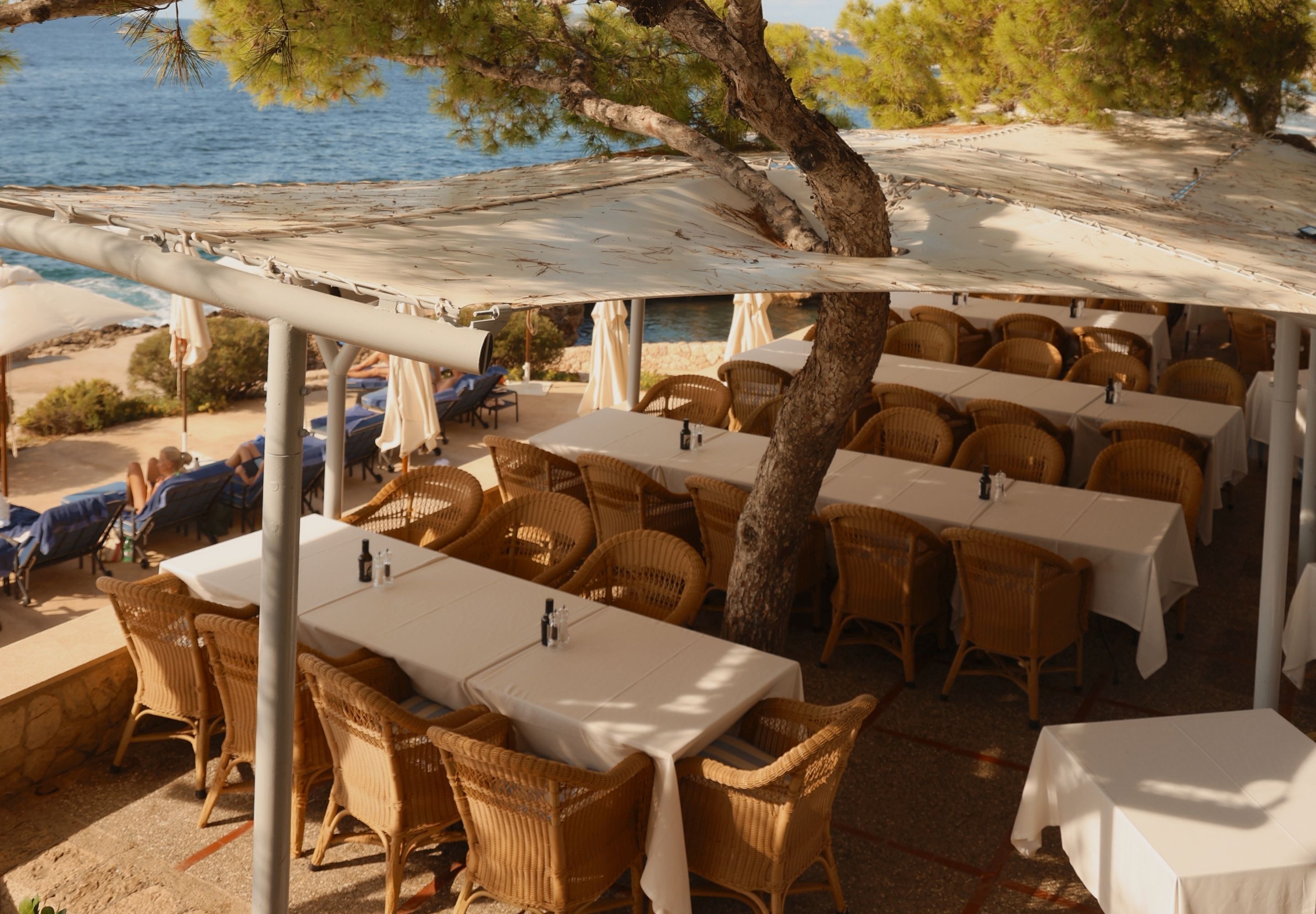 An outdoor dining area with wicker tables and chairs, a covered section, and a view of the sea.