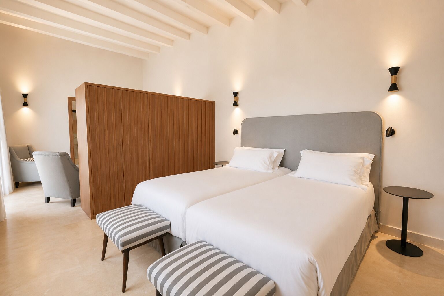 Een moderne hotelkamer met een groot bed, twee nachtkastjes, een houten garderobe, een grijze fauteuil en een gestreept voetenbankje.
