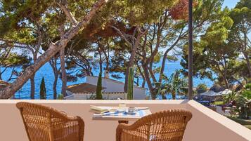 A balcony with wicker chairs, a table with a book and a cup, and a view of the sea and trees.