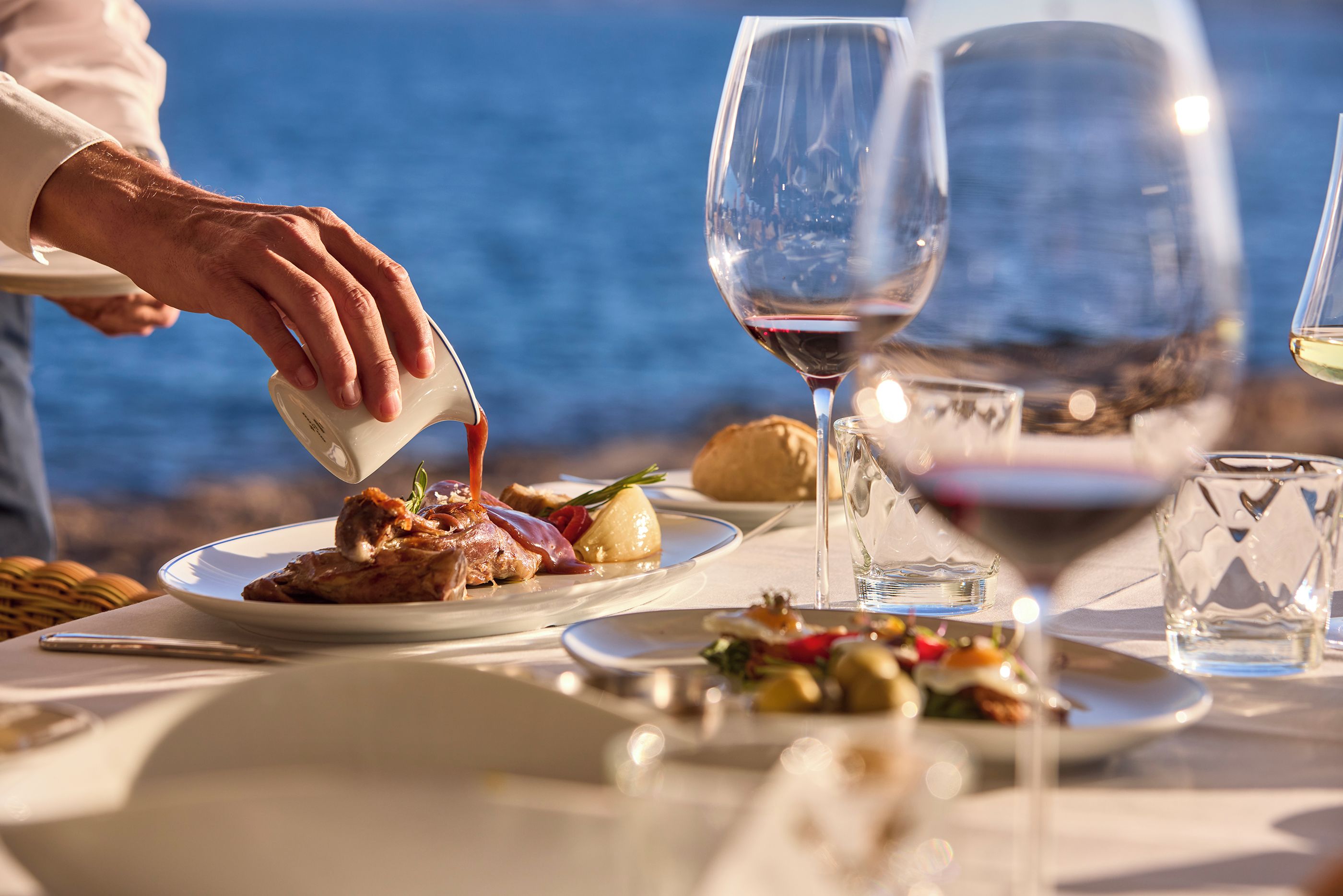 A dining table with plates of food, glasses of wine, and a person pouring a drink.