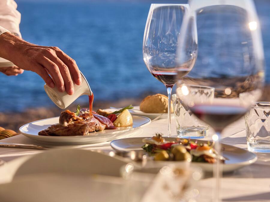 A dining table with plates of food, glasses of wine, and a person pouring a drink.