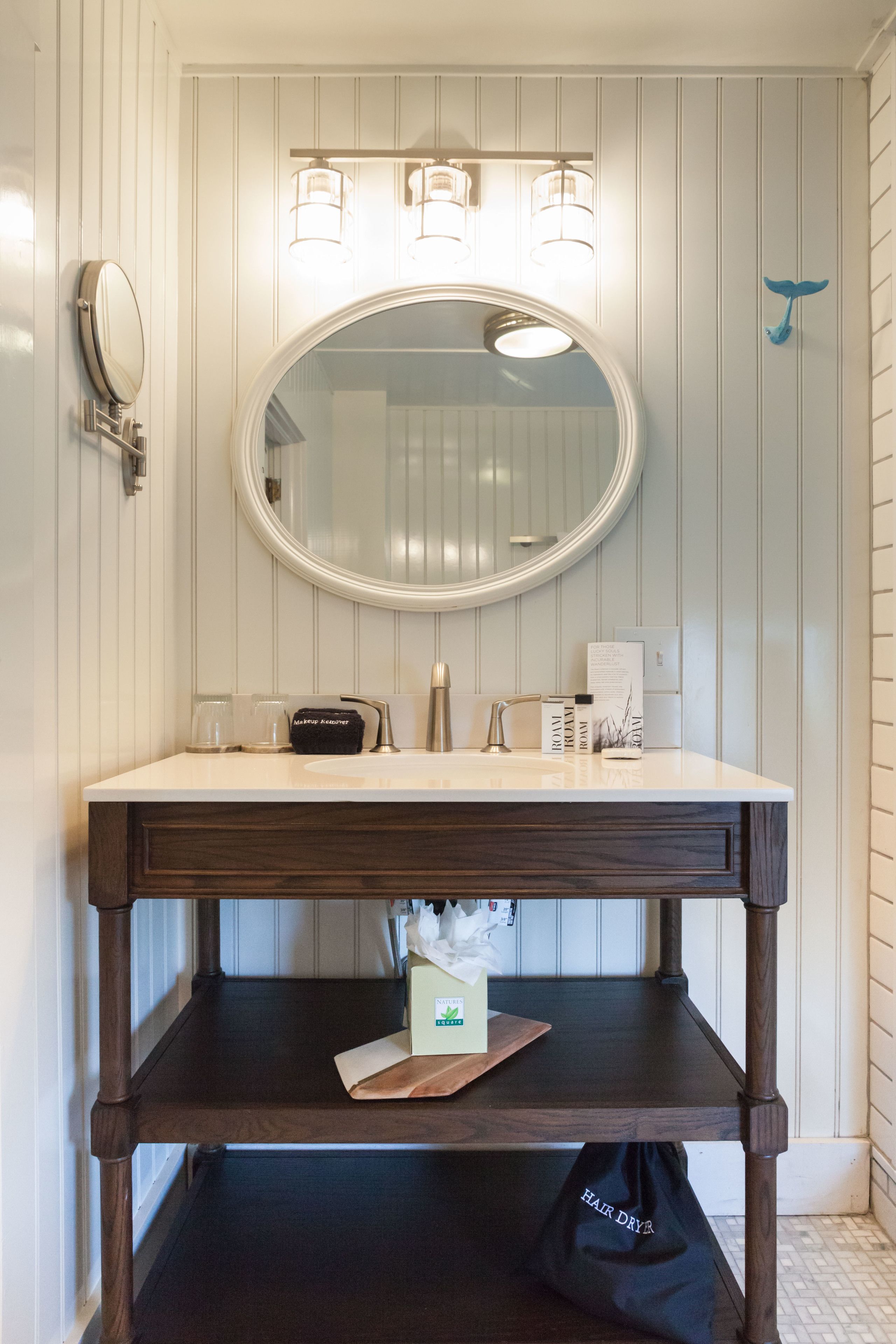 A bathroom with a round mirror, two wall-mounted lights, a vanity with a dark wooden base, and a hair dryer bag on the lower shelf.