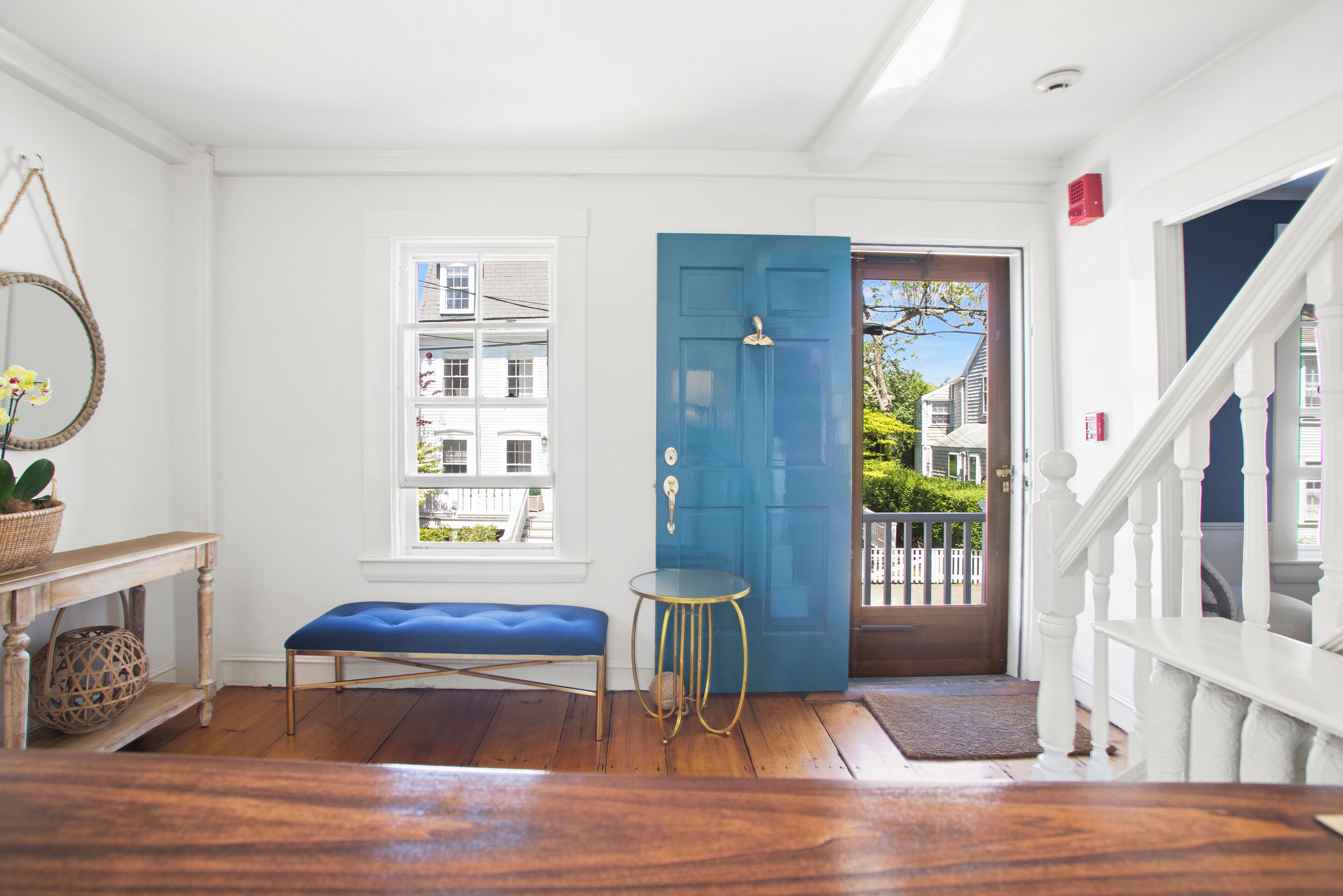 Un pasillo luminoso con una puerta azul, un banco, una mesita y una ventana que da a vistas de casas vecinas.