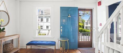 Un pasillo luminoso con una puerta azul, un banco, una mesita y una ventana que da a vistas de casas vecinas.