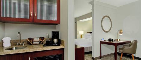 A hotel room with a kitchenette, including a sink, coffee maker, and a small refrigerator. There is a desk with a chair, a bedside table, and a lamp. The room has a carpeted floor and a mirror on the wall.