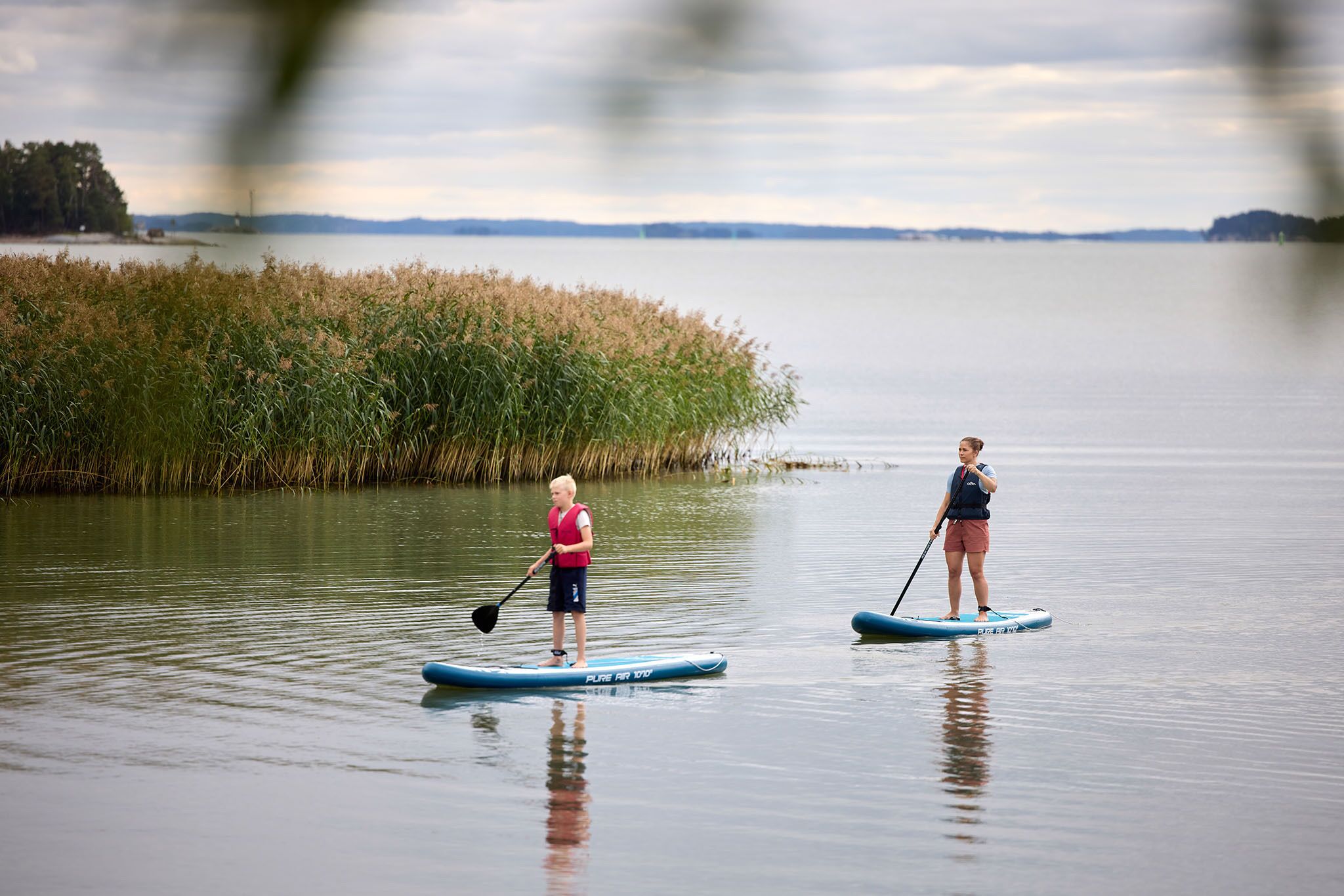 Duas pessoas praticando paddleboard em águas calmas, com uma linha costeira distante e vegetação.