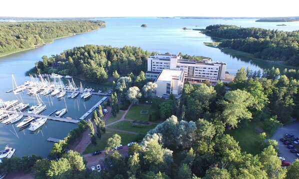 A marina with multiple boats docked, surrounded by dense forest and a large building with a modern design. - Ruissalo Spa Hotel (Turku)