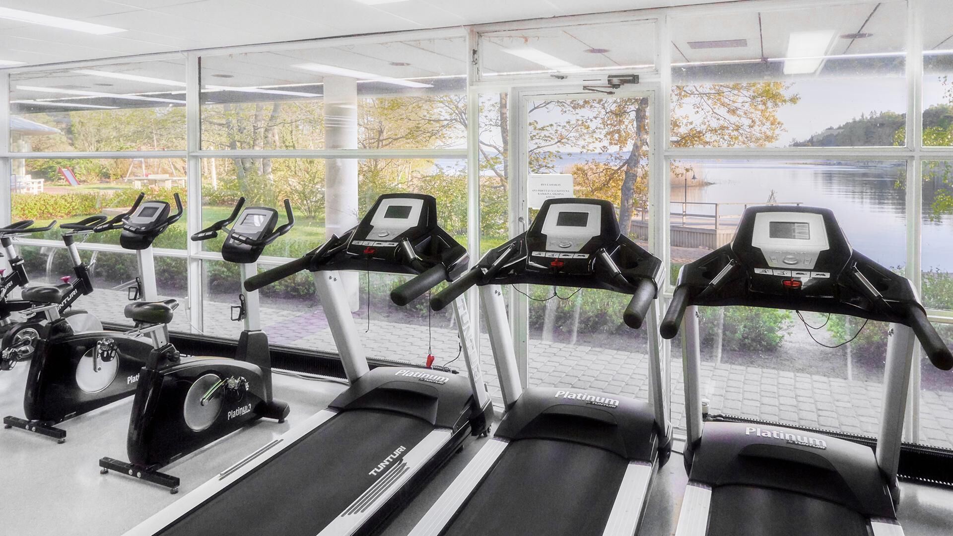 A gym with multiple treadmills and stationary bikes, large windows with a view of trees and a lake.