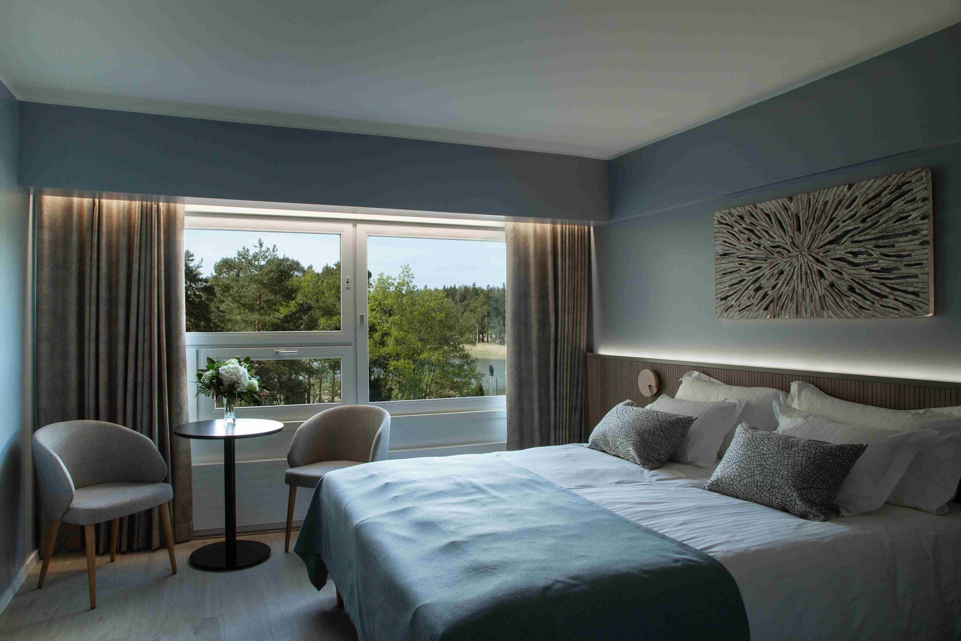 A hotel room with a bed, two chairs, a small table, a window with curtains, and a decorative wall piece.