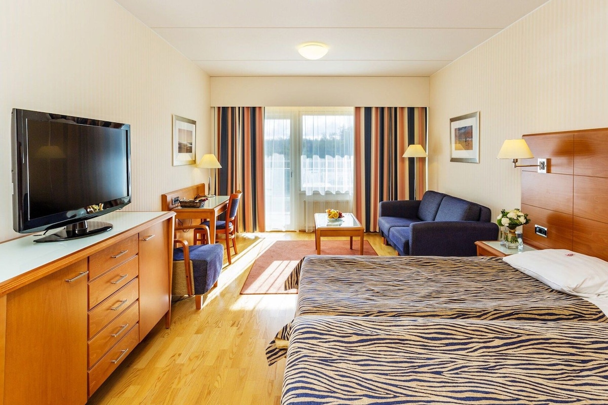 A hotel room with a bed, a TV, a desk, a chair, a sofa, and a window with curtains.