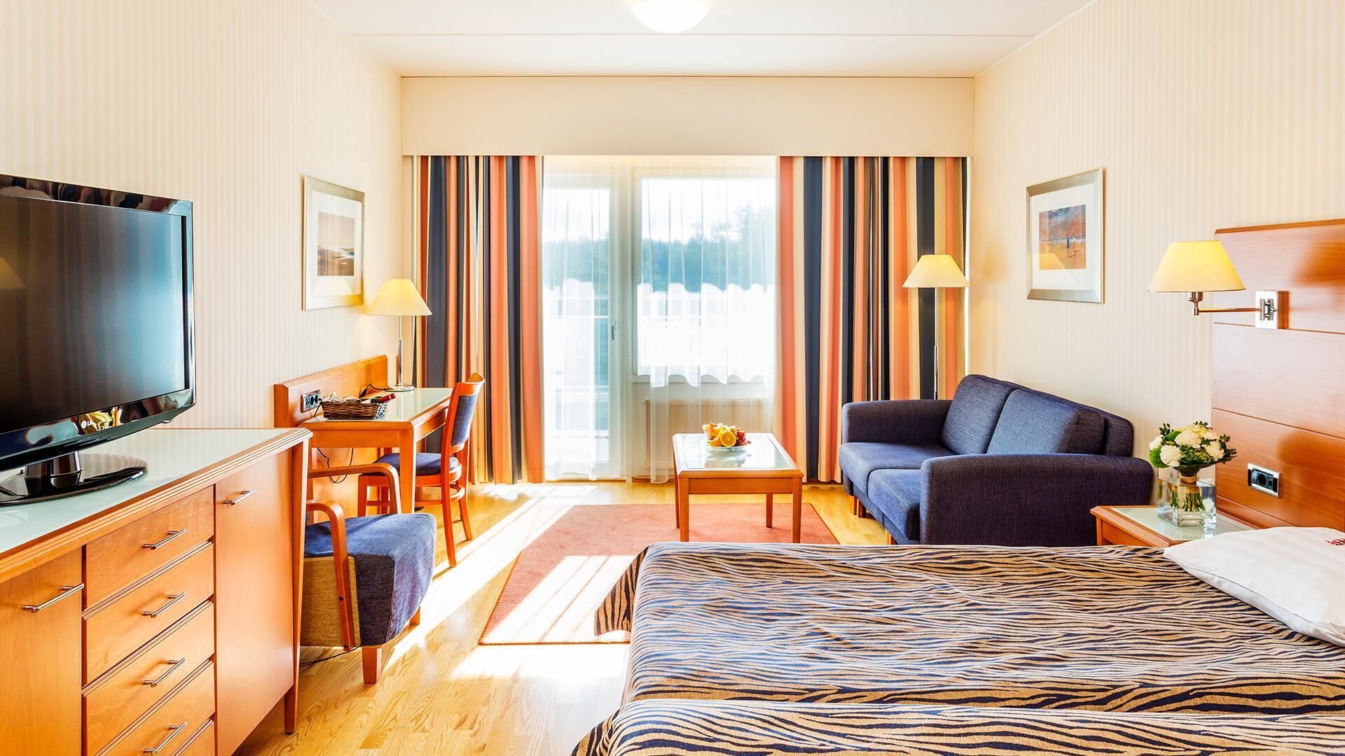 A hotel room with a bed, a television, a desk with a chair, a sofa, and a window with curtains.