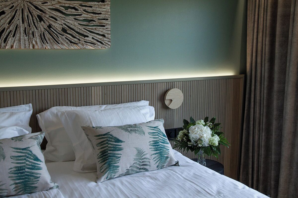 Un lit bien fait, avec une literie blanche et des coussins à motifs de fougères, un luminaire fixé au mur et un vase rempli de fleurs blanches.