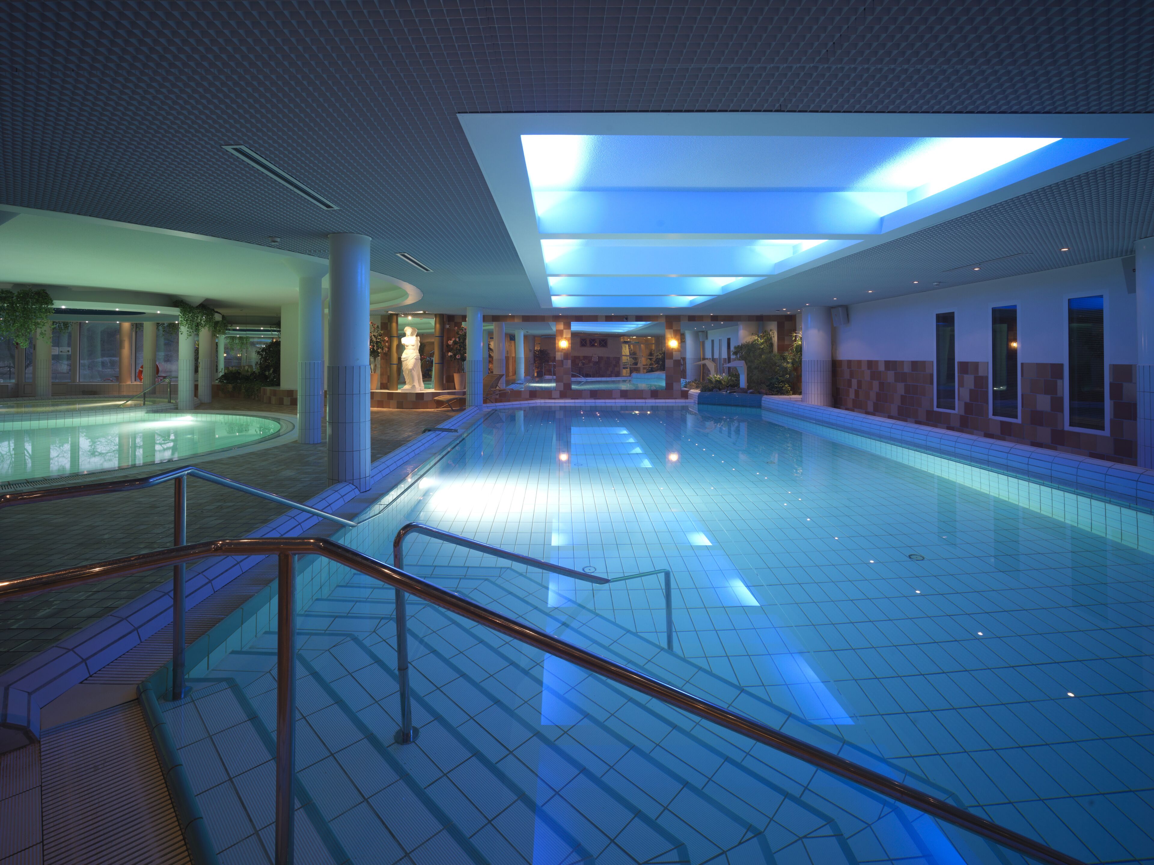 Una amplia piscina interior con suelo de baldosas, barandilla de vidrio y techo con tragaluces.