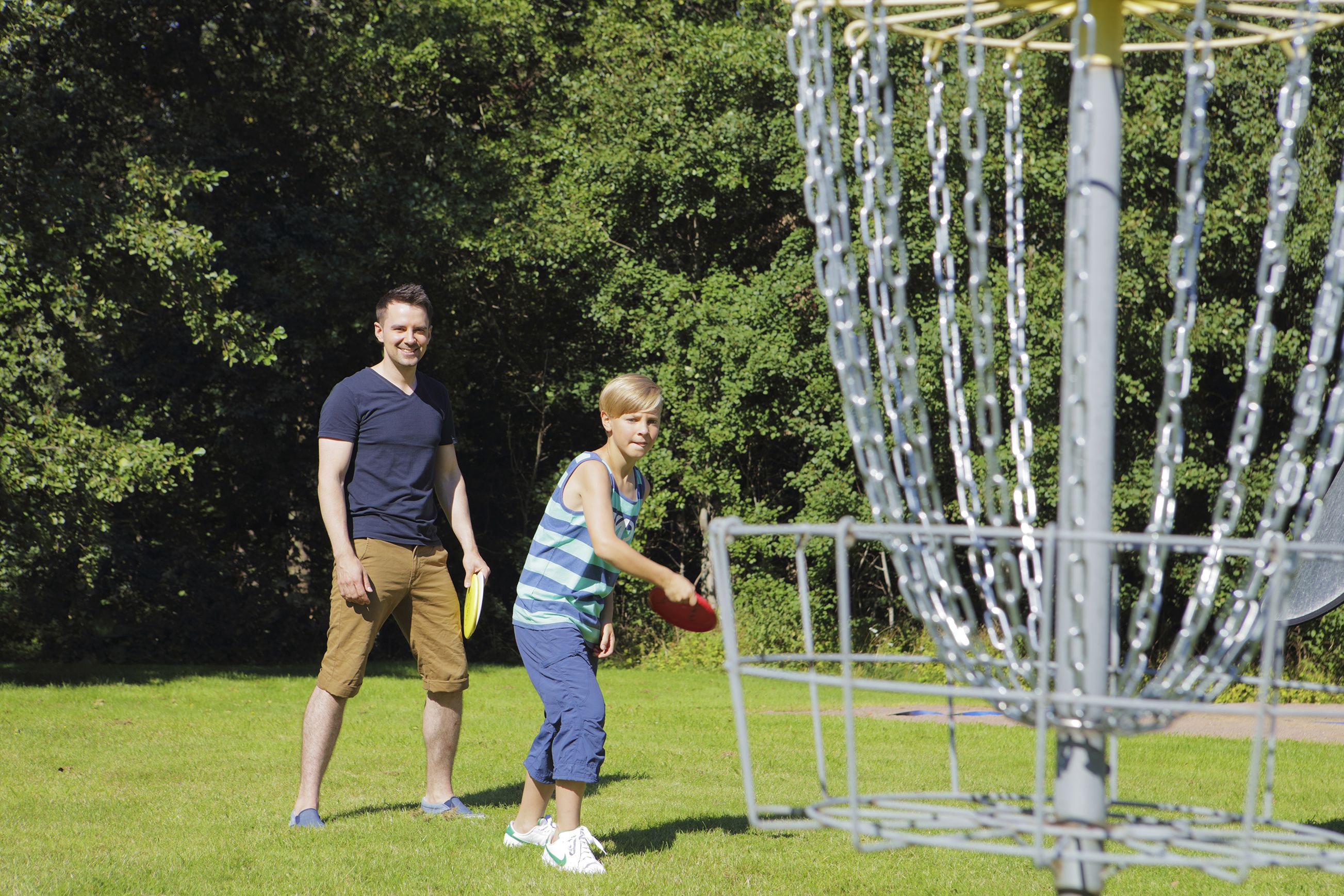 Two people playing frisbee in a park with a metal disc golf basket.