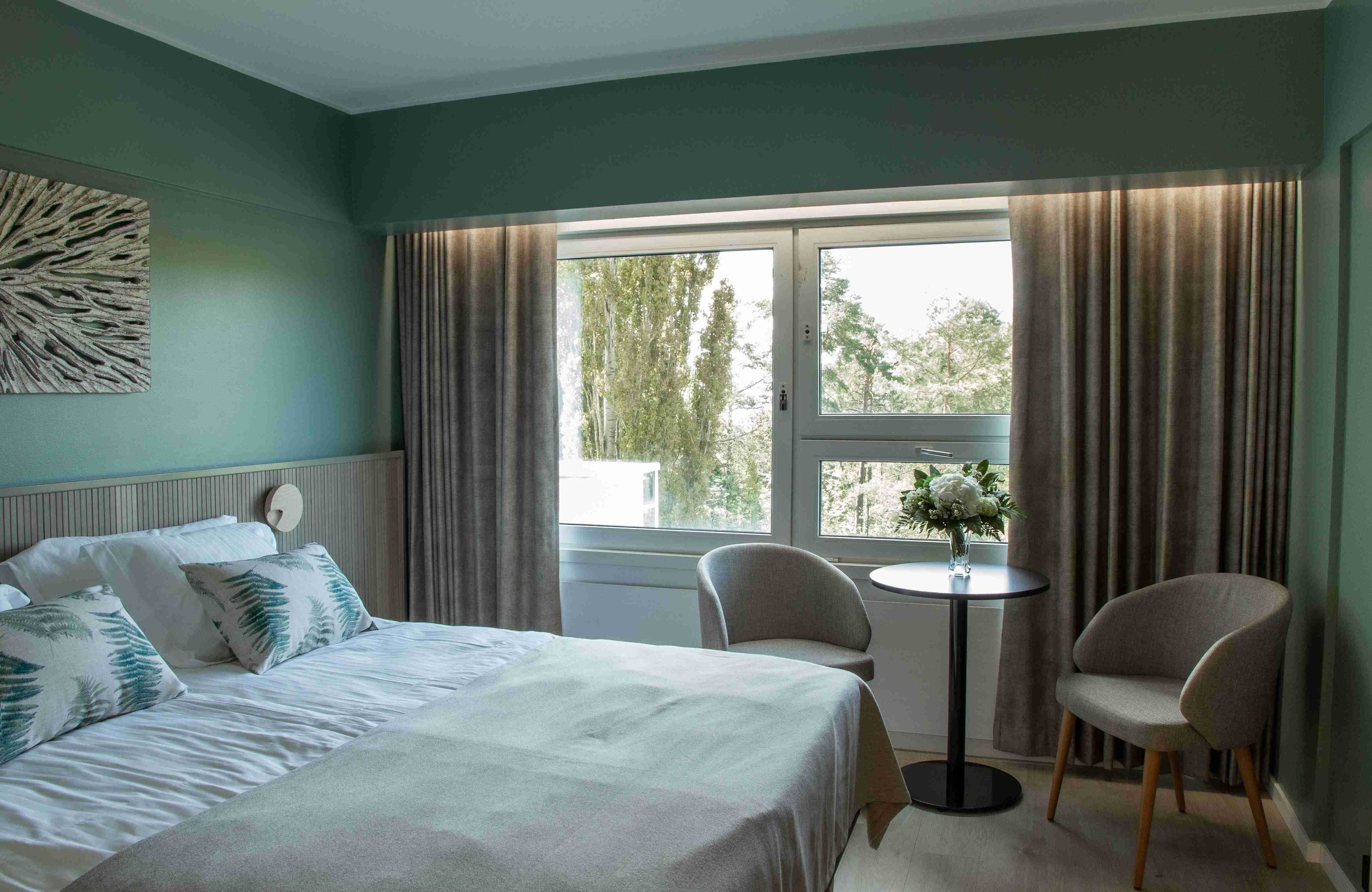 Ein Hotelzimmer mit einem Bett, zwei Stühlen, einem kleinen Tisch und einem Fenster mit Vorhängen.