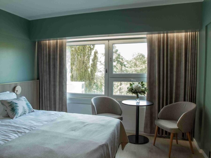 Ein Hotelzimmer mit einem Bett, zwei Stühlen, einem kleinen Tisch und einem Fenster mit Vorhängen.