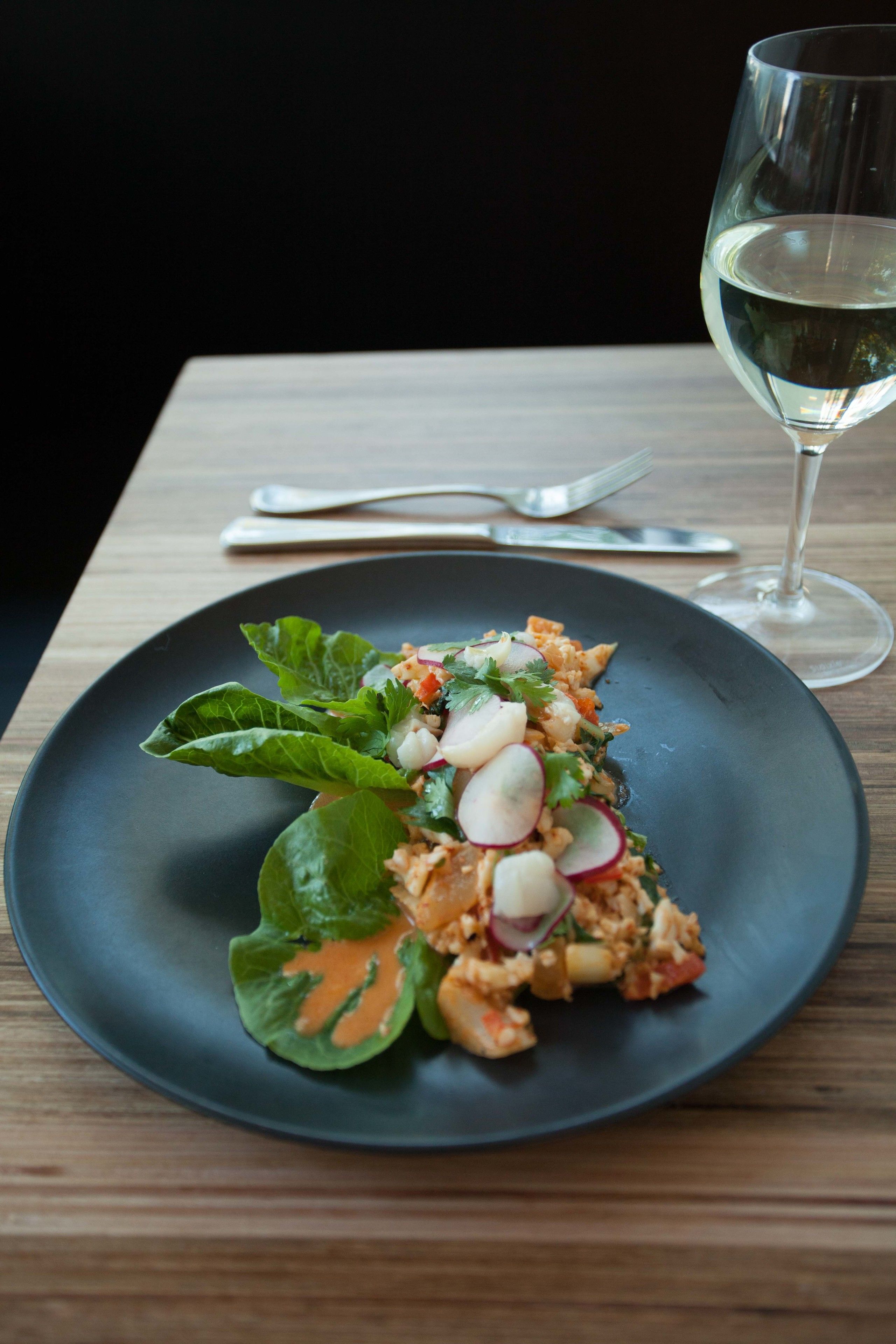 Un plato de comida sobre una mesa, con una copa de vino blanco y cubiertos.