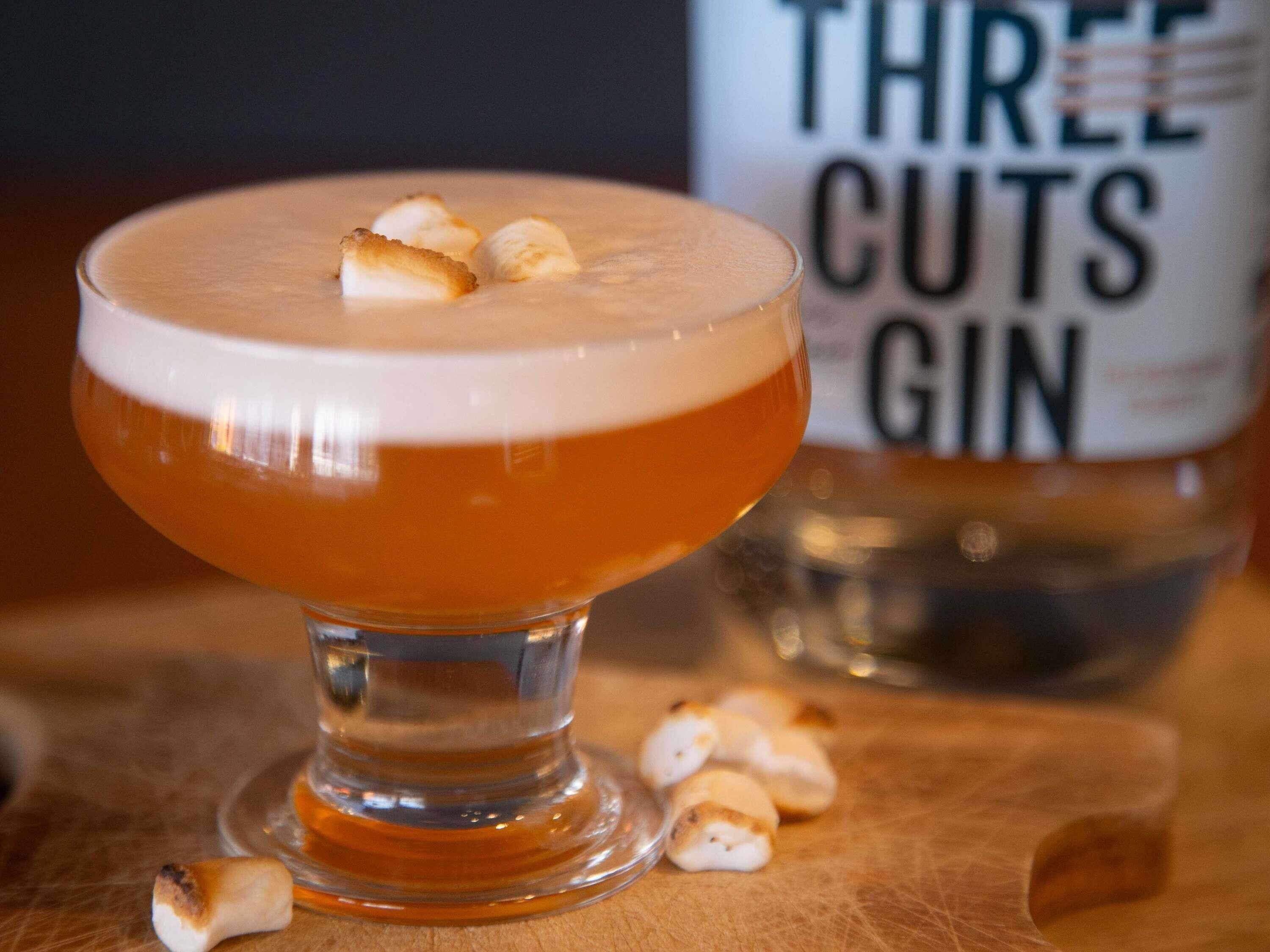 A cocktail in a stemmed glass with a foamy head, accompanied by toasted marshmallows on the rim.