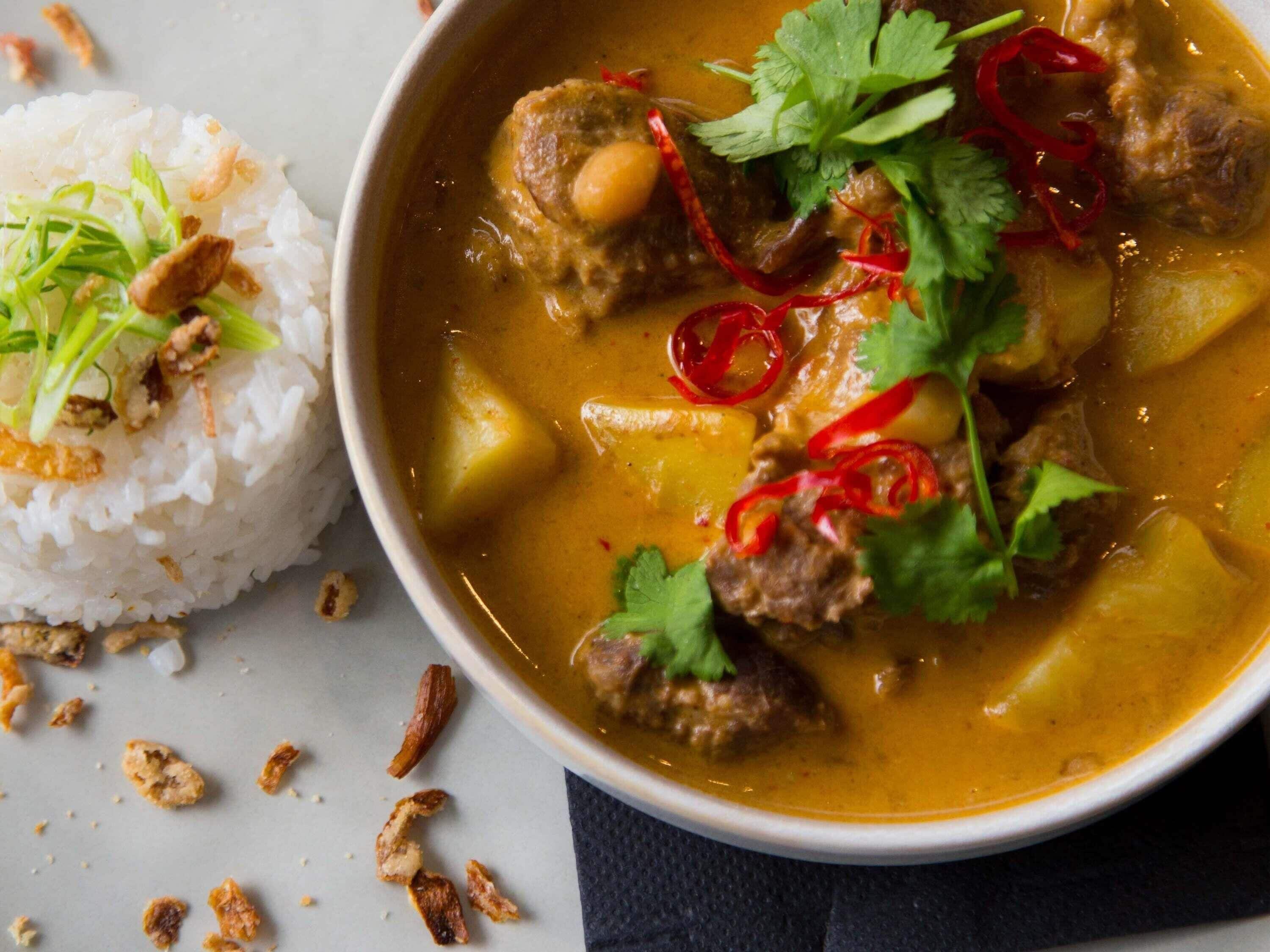 Uma tigela de curry com carne e vegetais, decorada com pimenta vermelha e coentro, acompanhada de arroz branco.