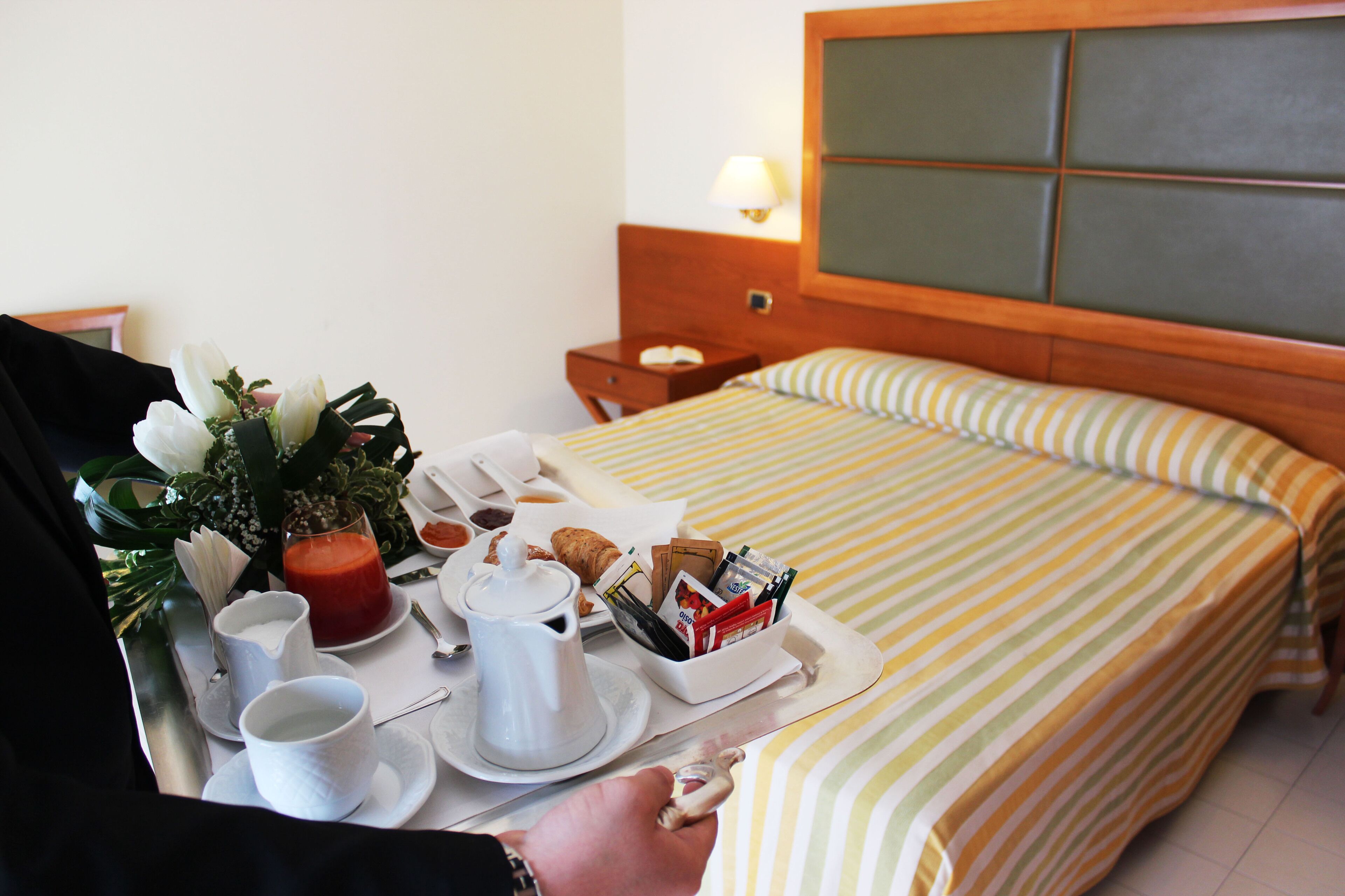 Una bandeja de desayuno bien dispuesta con tetera, tazas, pasteles y un pequeño jarrón de flores sobre una cama con ropa de cama a rayas y cabecera de madera.