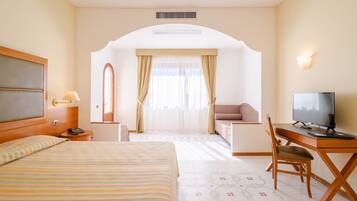 Quarto de hotel com cama, escrivaninha, televisão, poltrona, sofá, espelho e janela com cortinas.