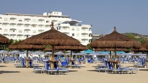 Une plage bordée de rangées de transats et d’ombrelles en chaume, un grand bâtiment blanc en arrière-plan et un paysage vallonné.