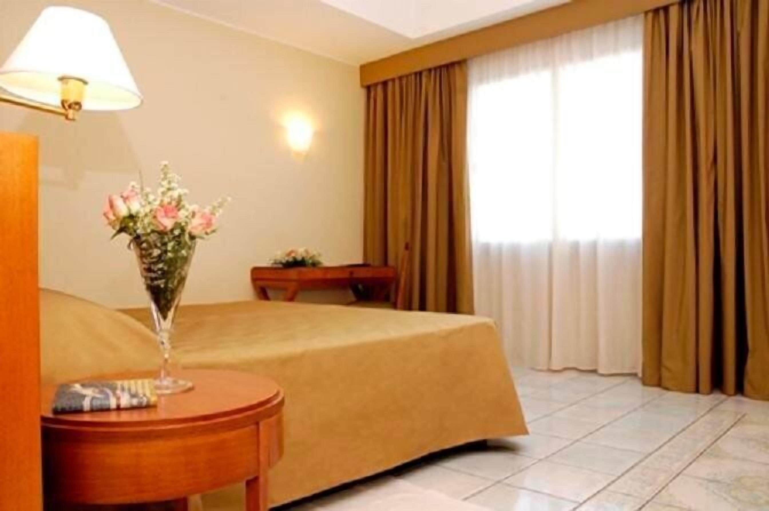 Ein Hotelzimmer mit einem Bett, einem Nachttisch mit einer Lampe und einer Blumenvase.