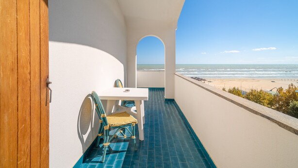 Un balcon avec une table blanche et des chaises en osier, donnant sur une plage et l’océan.