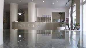 Un espace d’accueil doté d’un comptoir en verre, de plantes tropicales et d’une enseigne indiquant « Grand Hôtel Don Juan ».