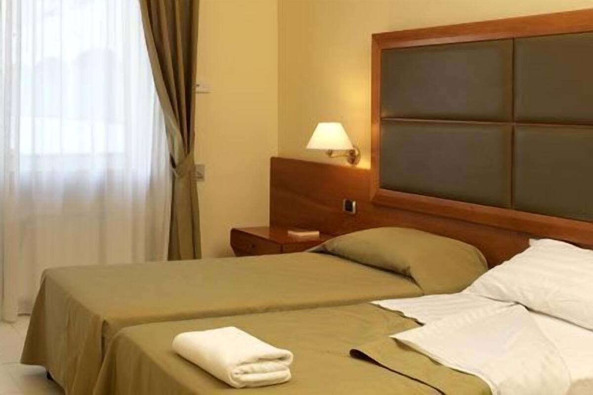 Habitación de hotel con una cama, un escritorio con computadora, una mesita de noche con lámpara y una ventana con cortinas.