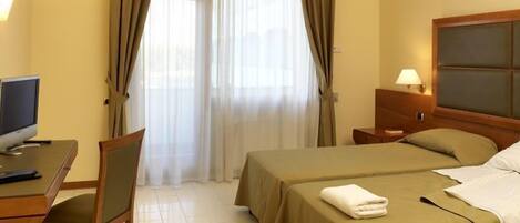 A hotel room with a bed, a desk with a computer, a nightstand with a lamp, and a window with curtains.