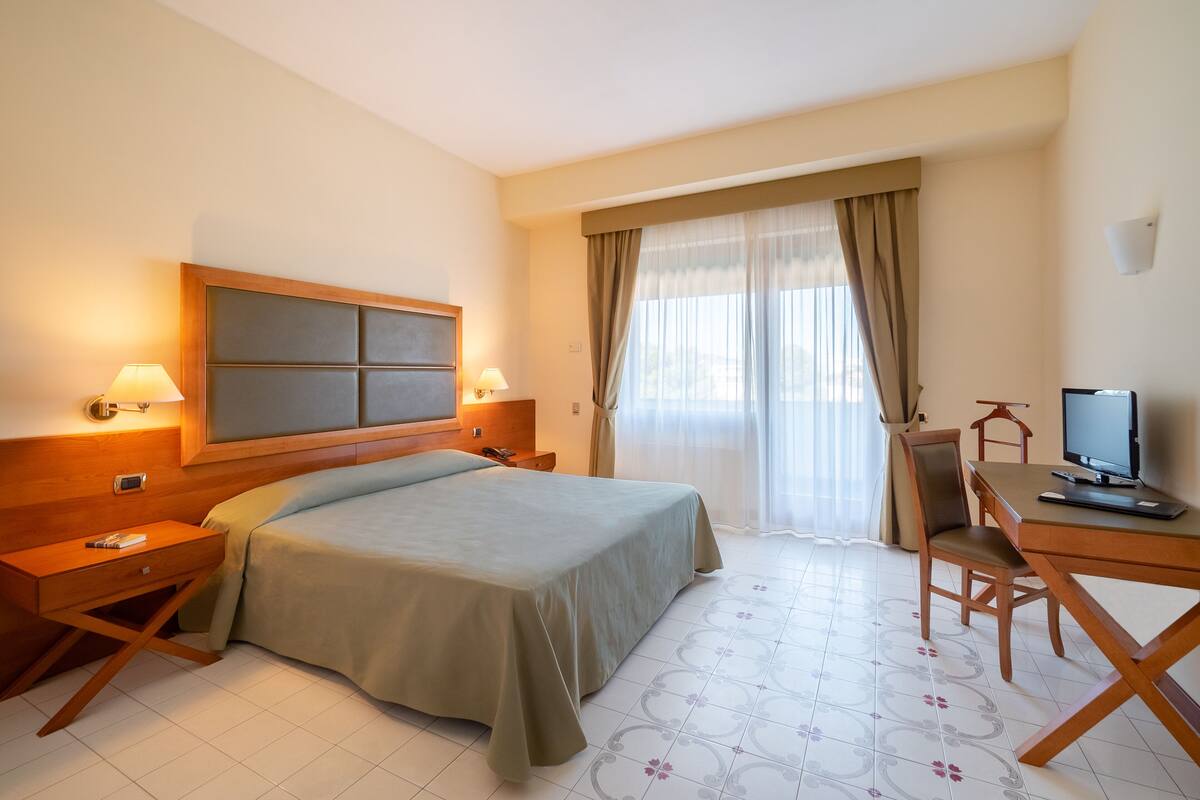 Una habitación de hotel con cama, mesitas de noche, un escritorio con silla, televisor y ventana con cortinas.