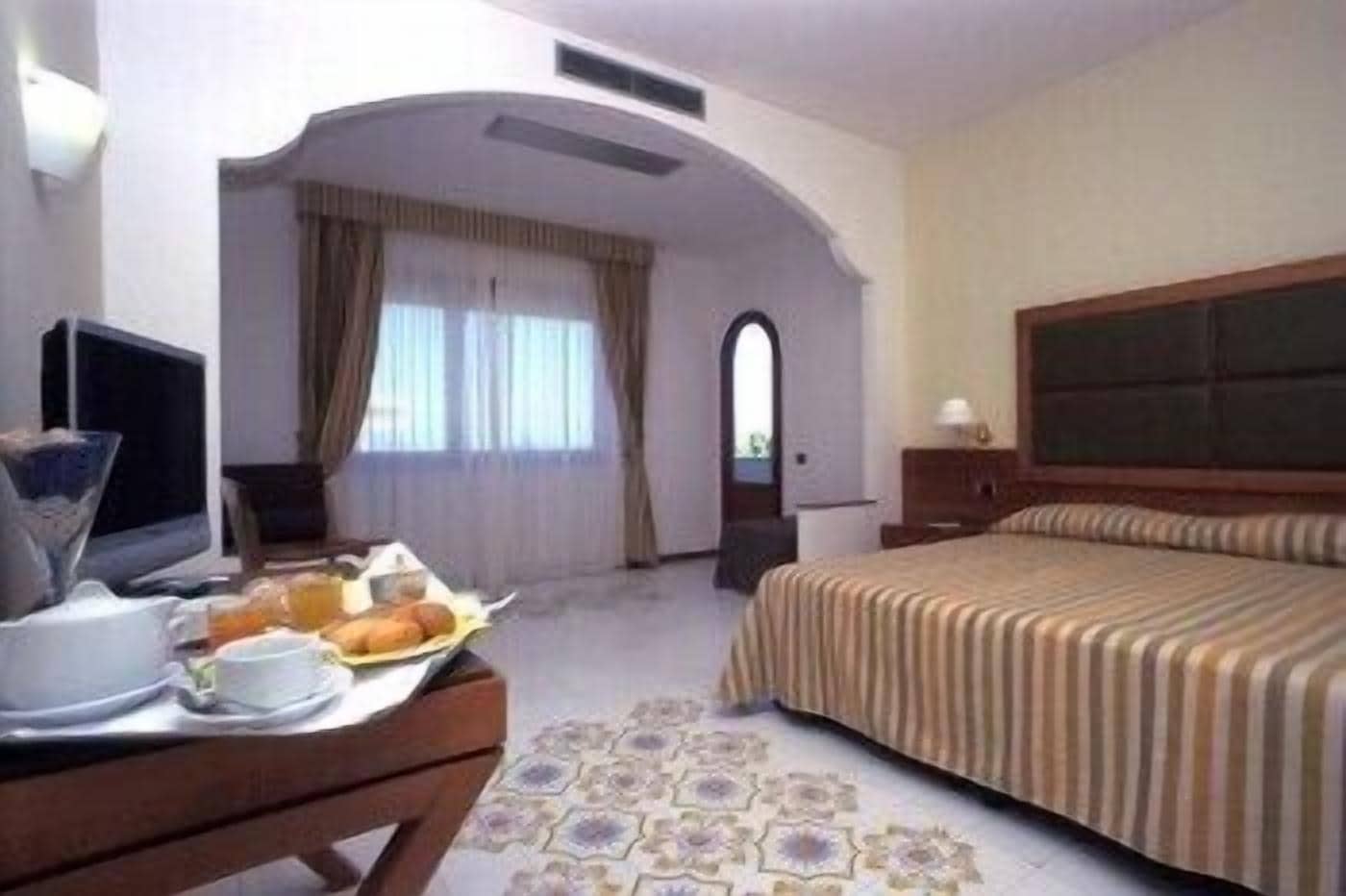A hotel room with a bed, a desk with breakfast items, a television, and a window with curtains.